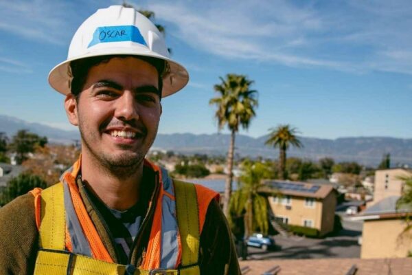 Oscar Flores on rooftop during solar installation, smiling at camera. View of a residential neighborhood behind him with palm trees.
